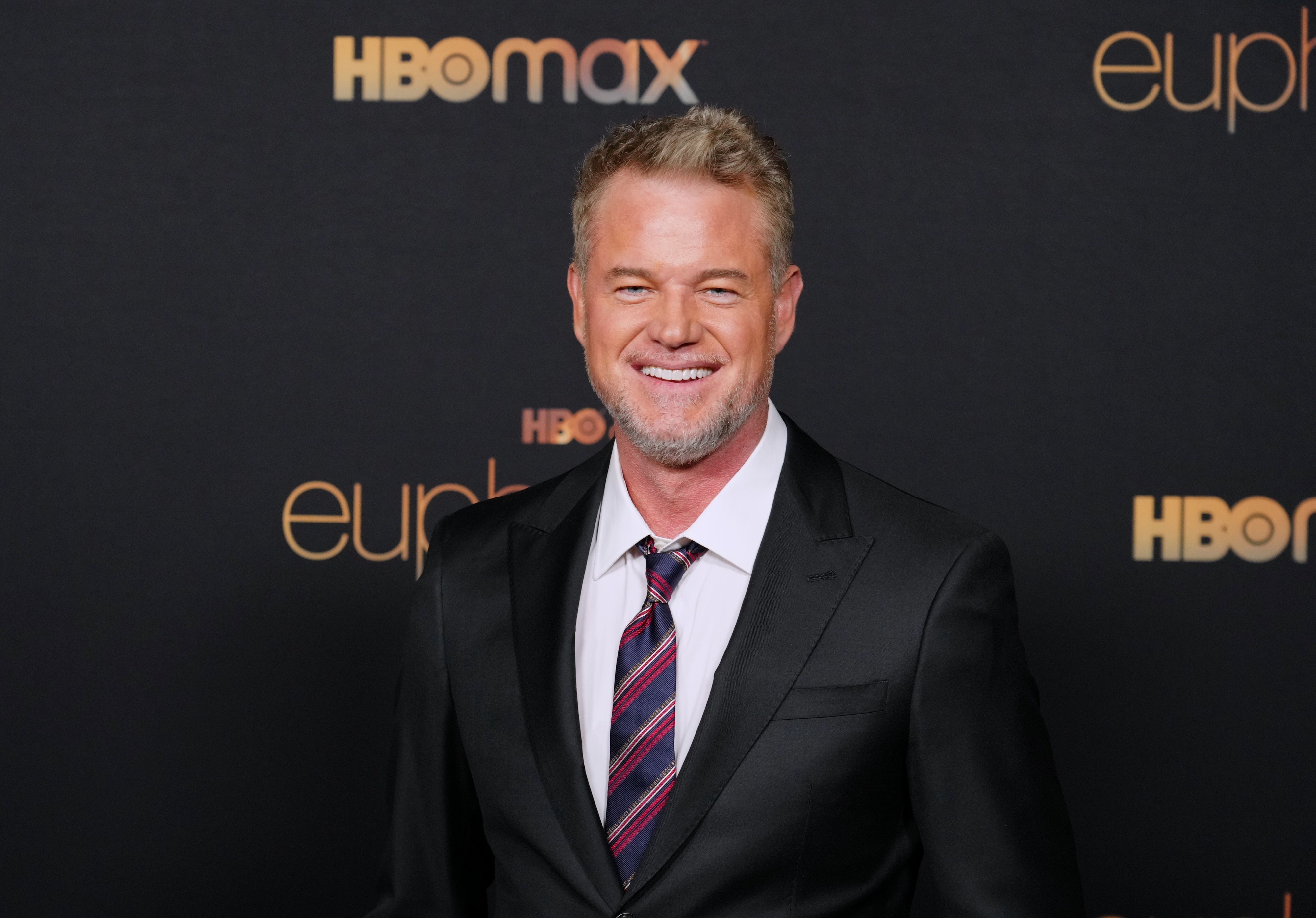 Eric Dane stands smiling on the red carpet at the HBO Max and Euphoria event, wearing a dark suit, white shirt, and striped tie