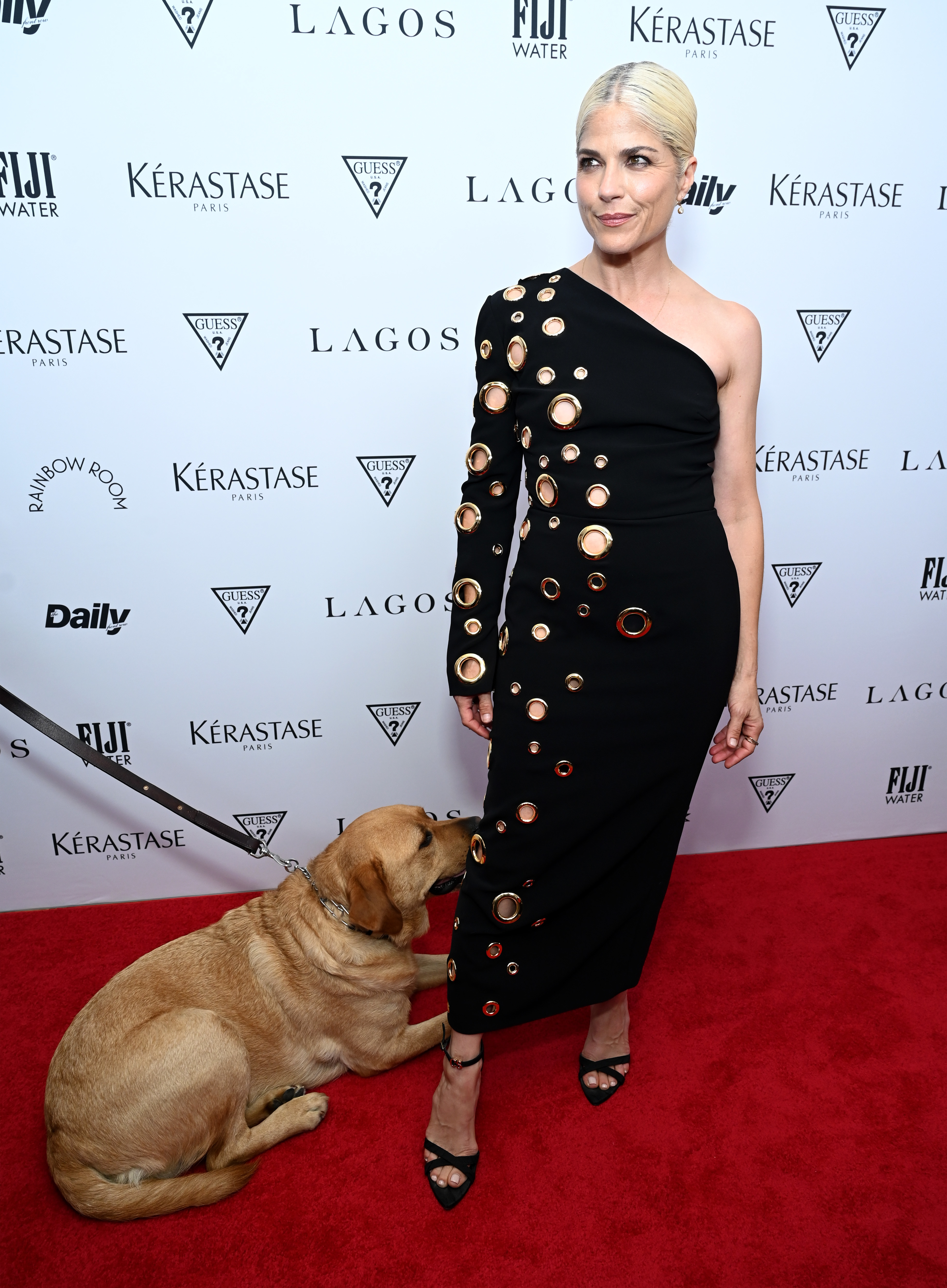 Selma Blair on the red carpet in an asymmetrical dress with circular cutouts, standing with her service dog. Background displays various logos