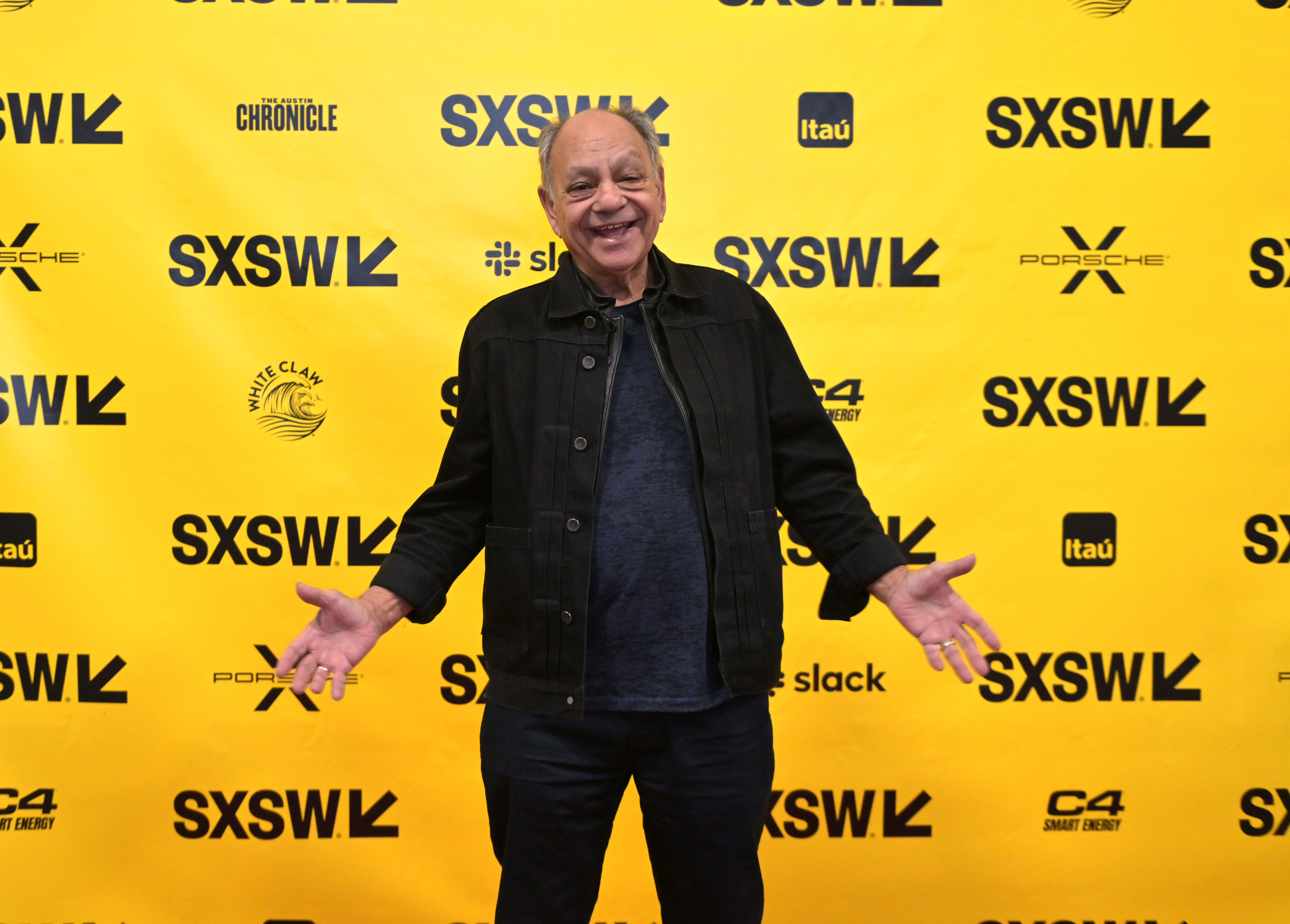 Cheech Marin stands on a yellow SXSW backdrop, smiling with arms open, wearing a black jacket and dark pants