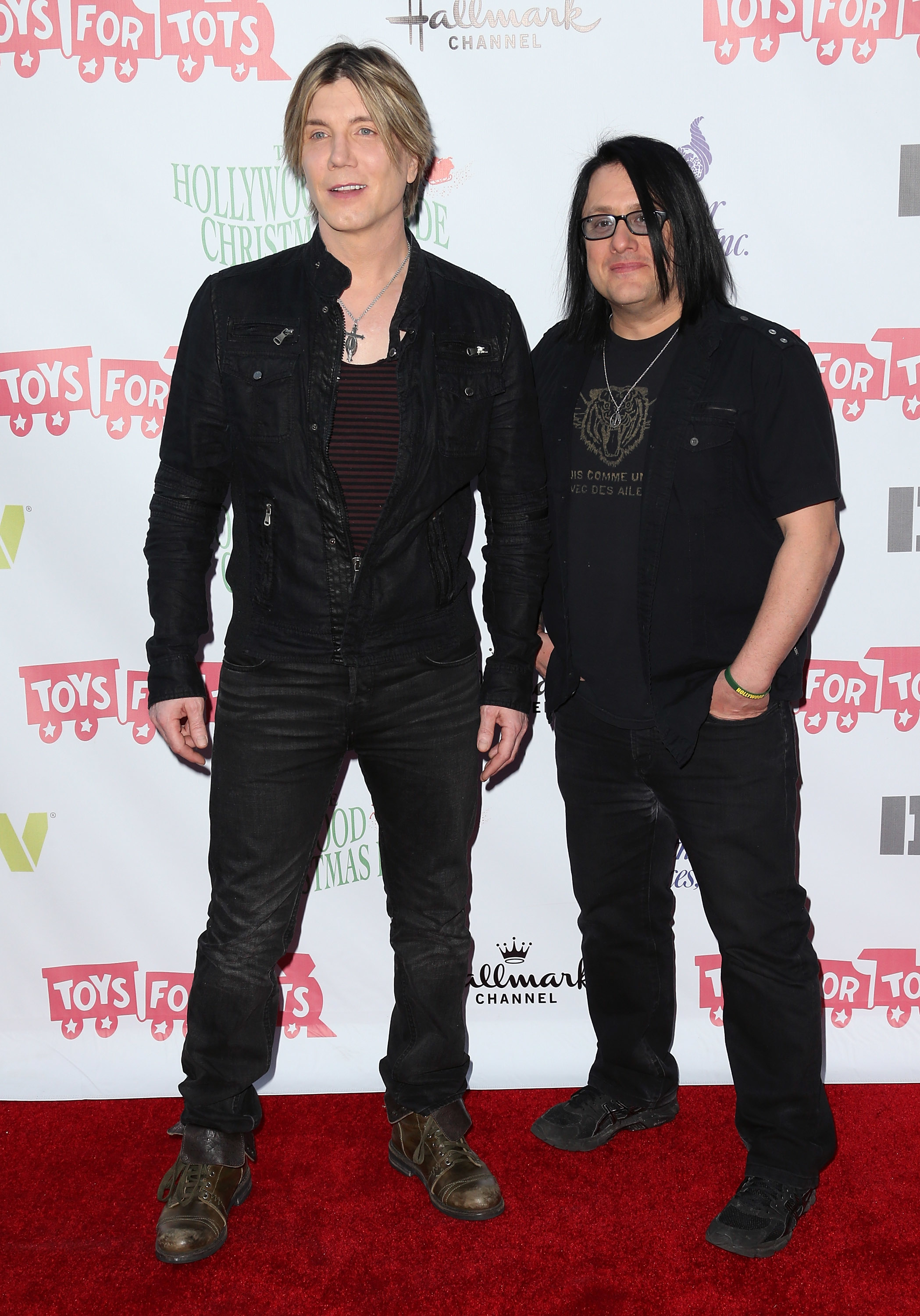 Two men, one with light hair in black jacket and pants, the other with dark hair in a black shirt, stand on a red carpet at the Toys for Tots event