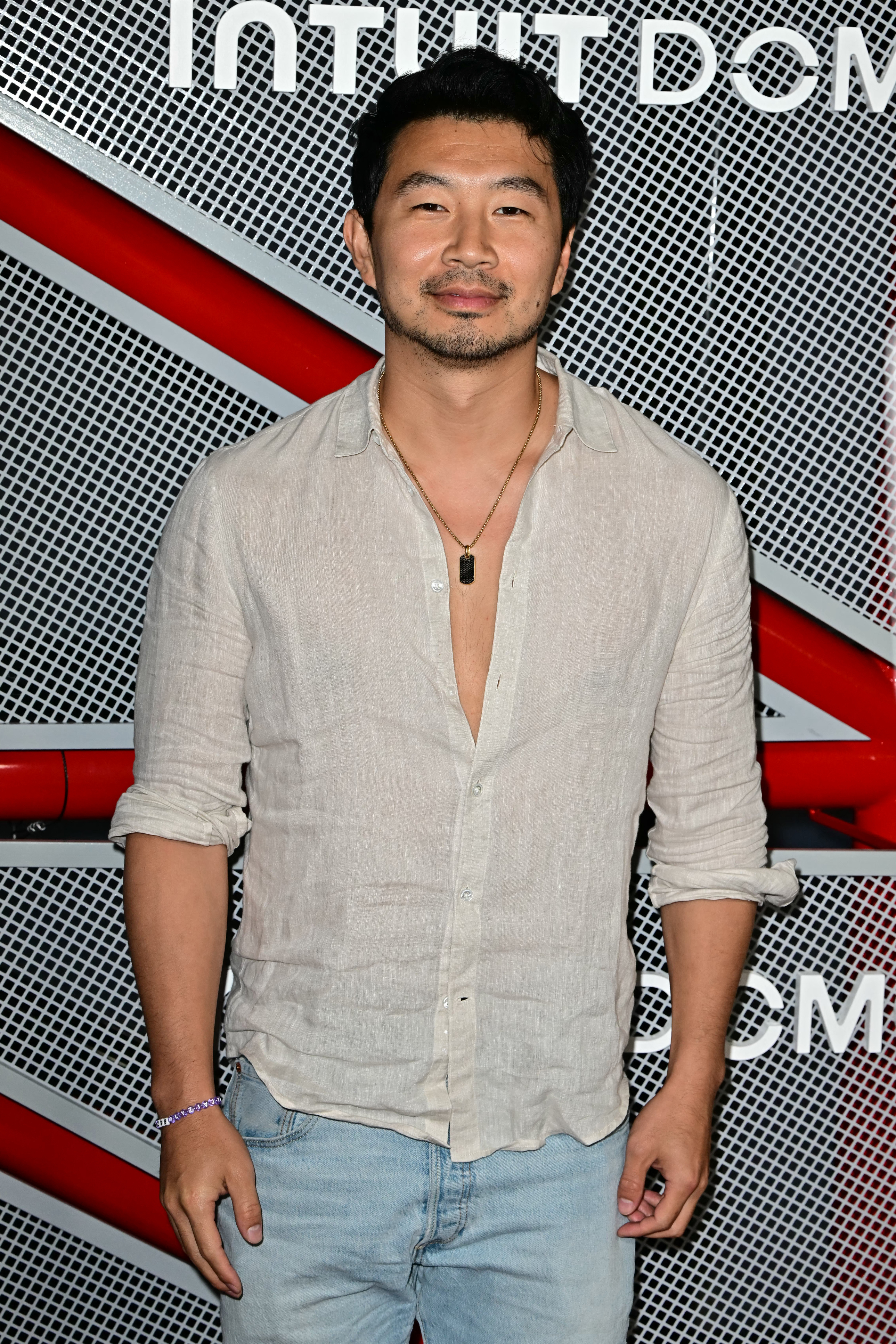 Simu Liu stands in front of an Intuit Dome backdrop, wearing a light, casual button-up shirt and light blue jeans