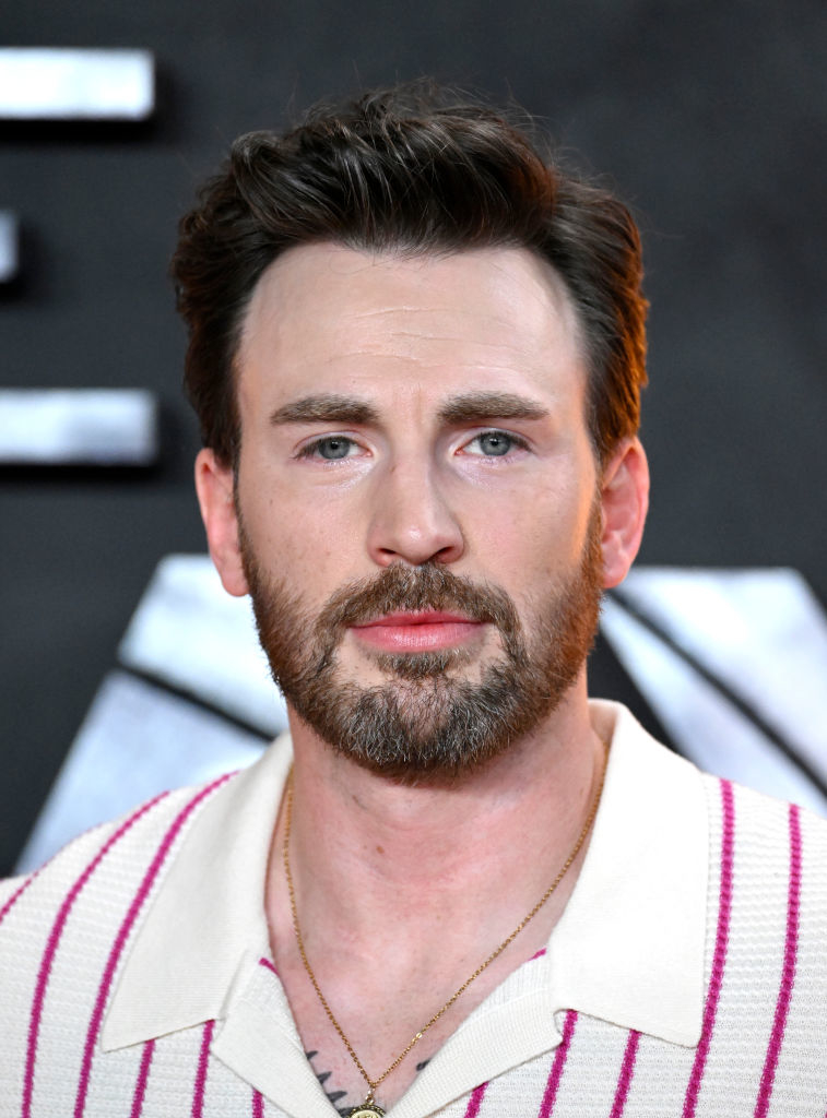 Chris Evans at a public event, wearing a half-buttoned white collared shirt with pink stripes, smiling gently at the camera