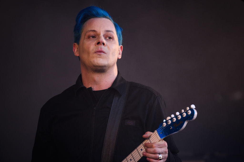 Jack White playing a guitar on stage, wearing a button-up shirt and sporting blue hair