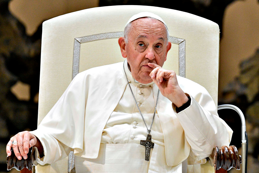 Pope Francis sits in a chair, wearing his traditional white papal attire and a cross necklace, with his hand resting near his mouth