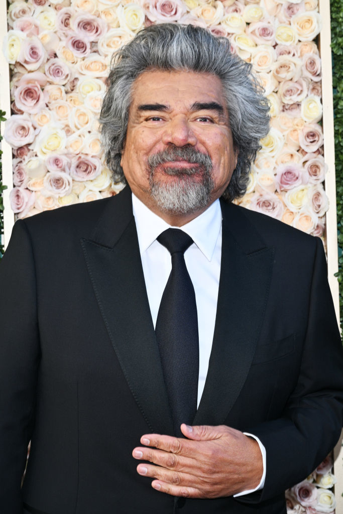 George Lopez on a floral background, wearing a classic black suit and tie, smiling with his hand on his stomach