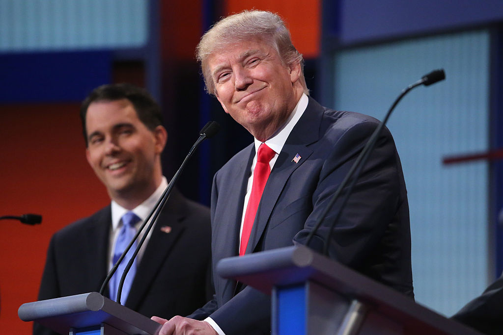 Donald Trump smiles at the debate podium, wearing a suit and tie, with another man standing behind another podium who is also smiling