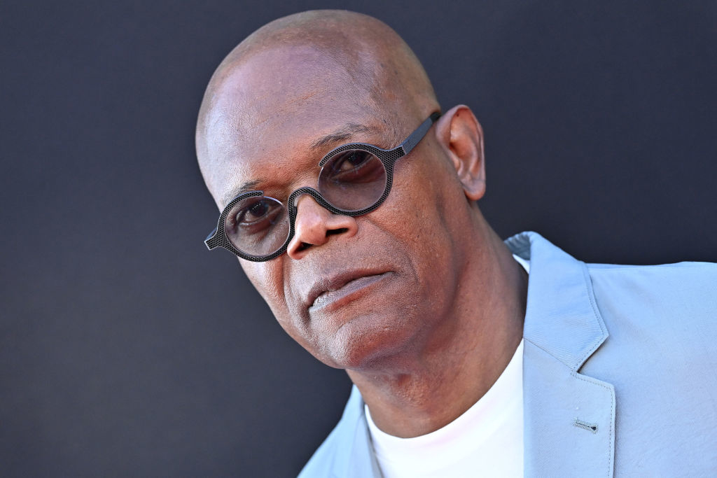 Samuel L. Jackson wears a light-colored blazer and a white shirt, paired with round sunglasses, posing against a dark background