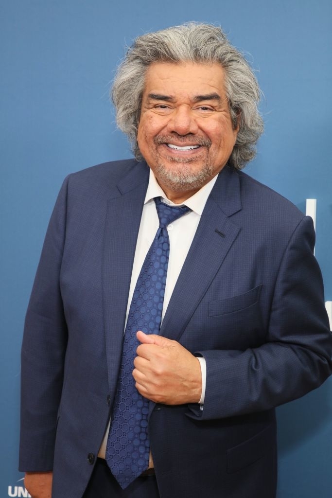 George Lopez smiling and wearing a suit with a tie, in front of a blue background at a media event