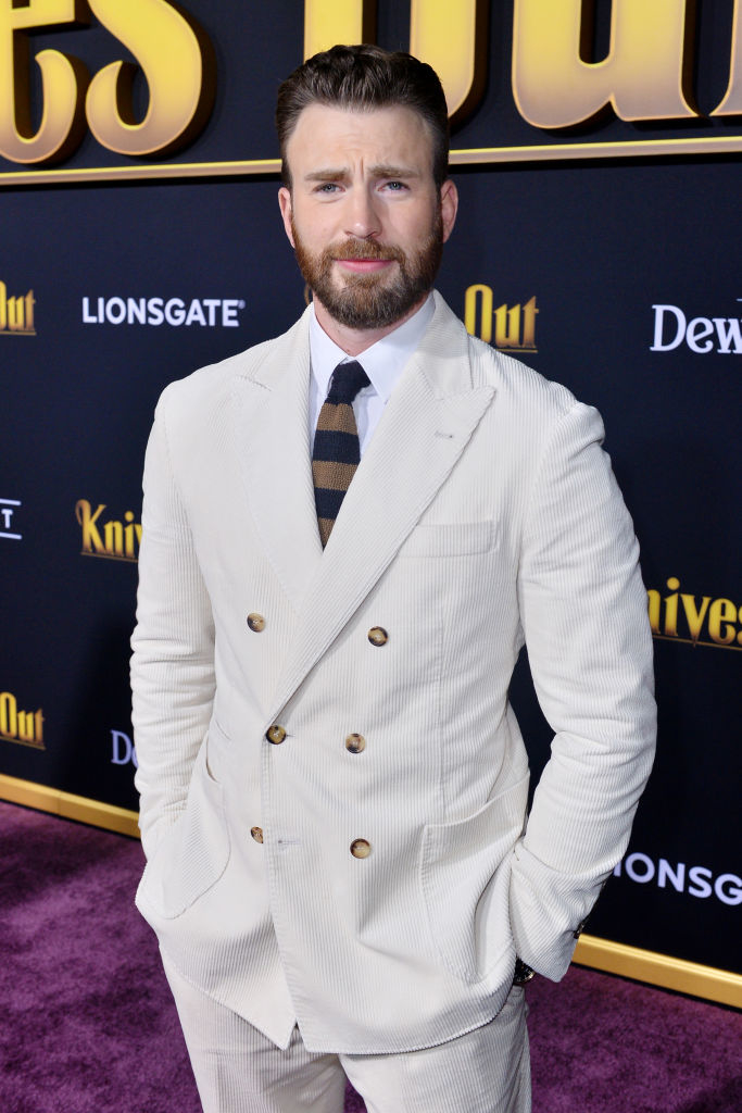 Chris Evans on the red carpet at a Knives Out event, wearing a light-colored double-breasted suit with a striped tie