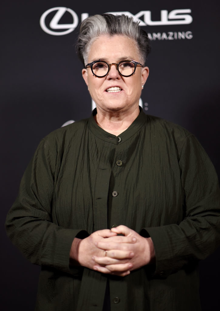 Rosie O&#x27;Donnell stands in front of a black backdrop with a Lexus logo, wearing a dark buttoned jacket and glasses. She has short, styled hair