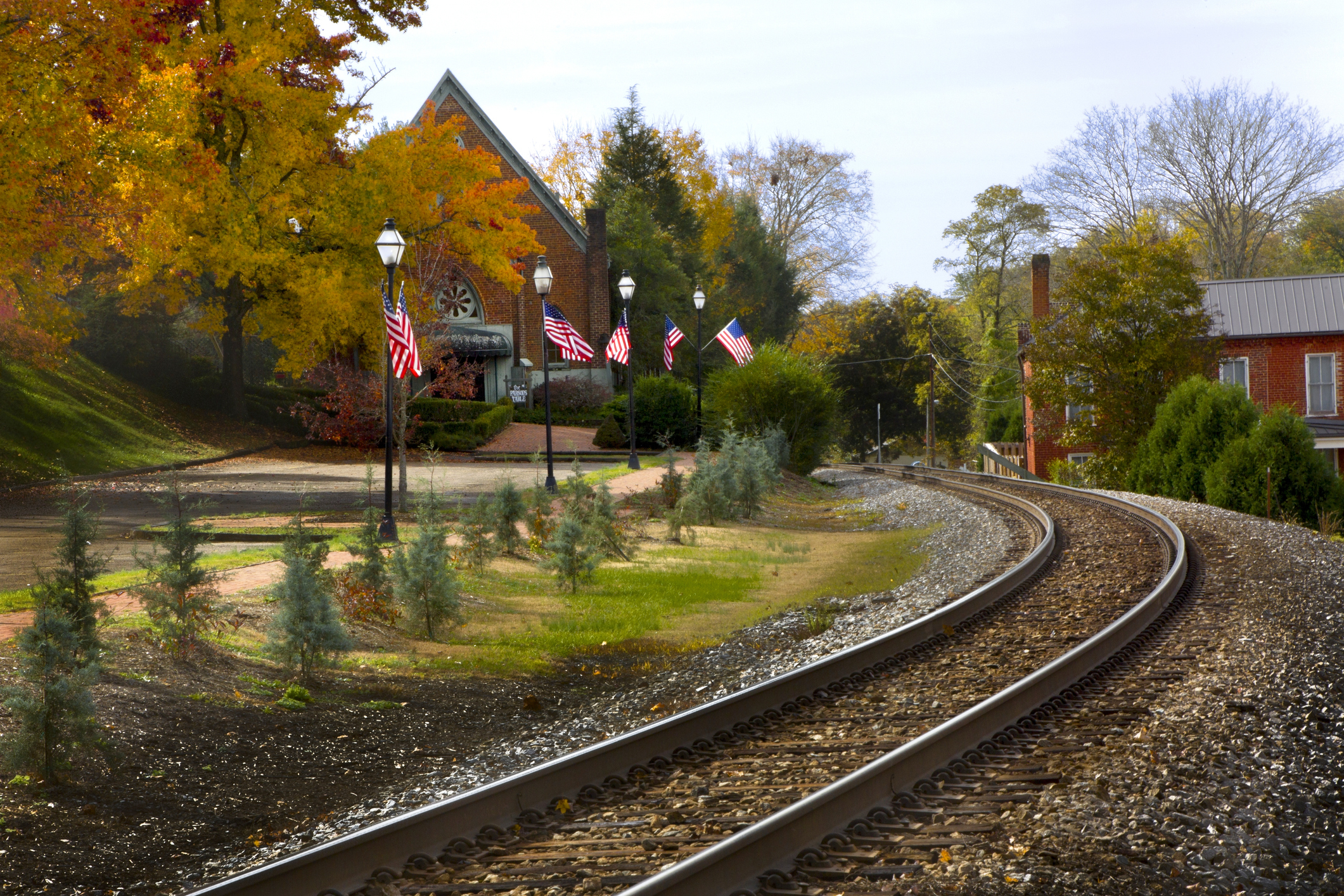 A curved railroad track leads to a quaint town with trees, a brick building, and American flags lining the path. The scene suggests a peaceful, small-town setting