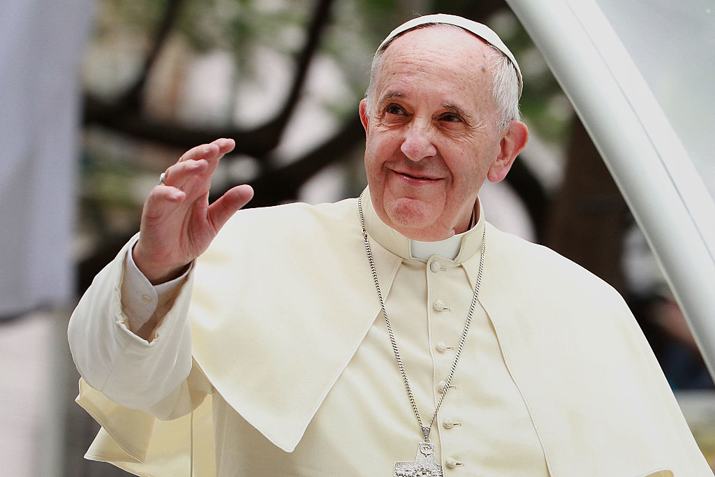 Pope Francis wears white clerical attire with a silver cross necklace, smiling and waving during a public event