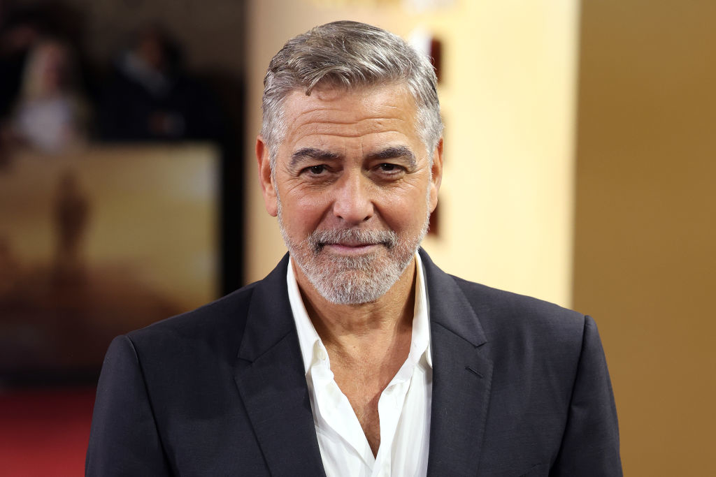 George Clooney at a red carpet event, wearing a dark blazer over a white shirt, smiling at the camera