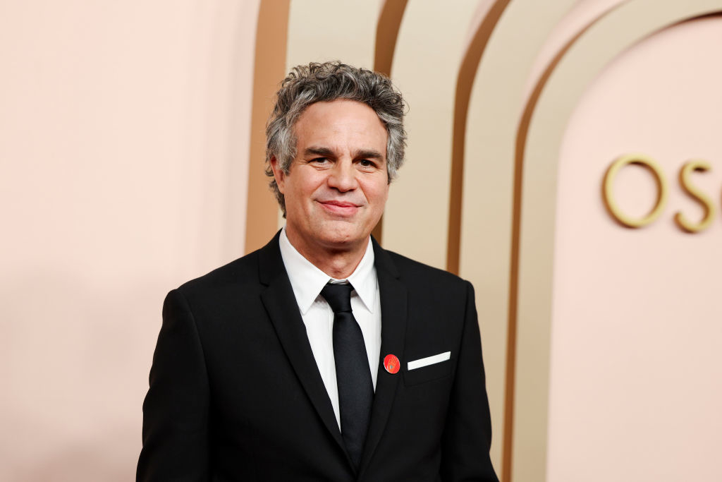 Mark Ruffalo stands on the red carpet, wearing a black suit and tie with a red pin on his left lapel. The backdrop features light-colored arches