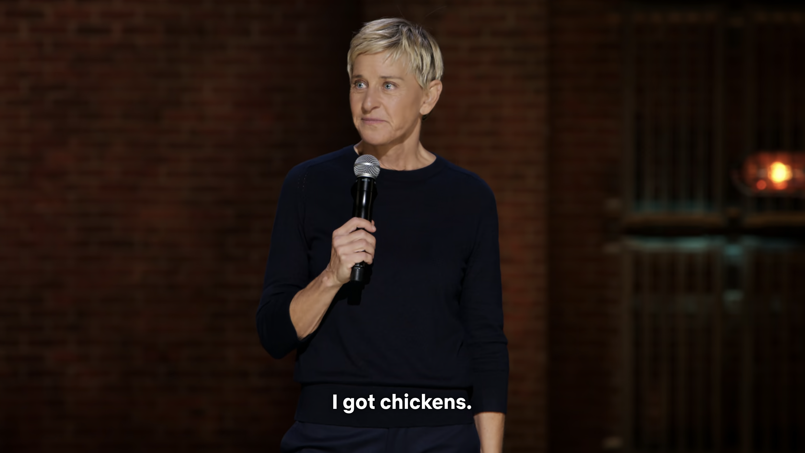 Ellen DeGeneres, wearing a simple outfit, stands on stage holding a microphone with the caption &quot;I got chickens&quot;