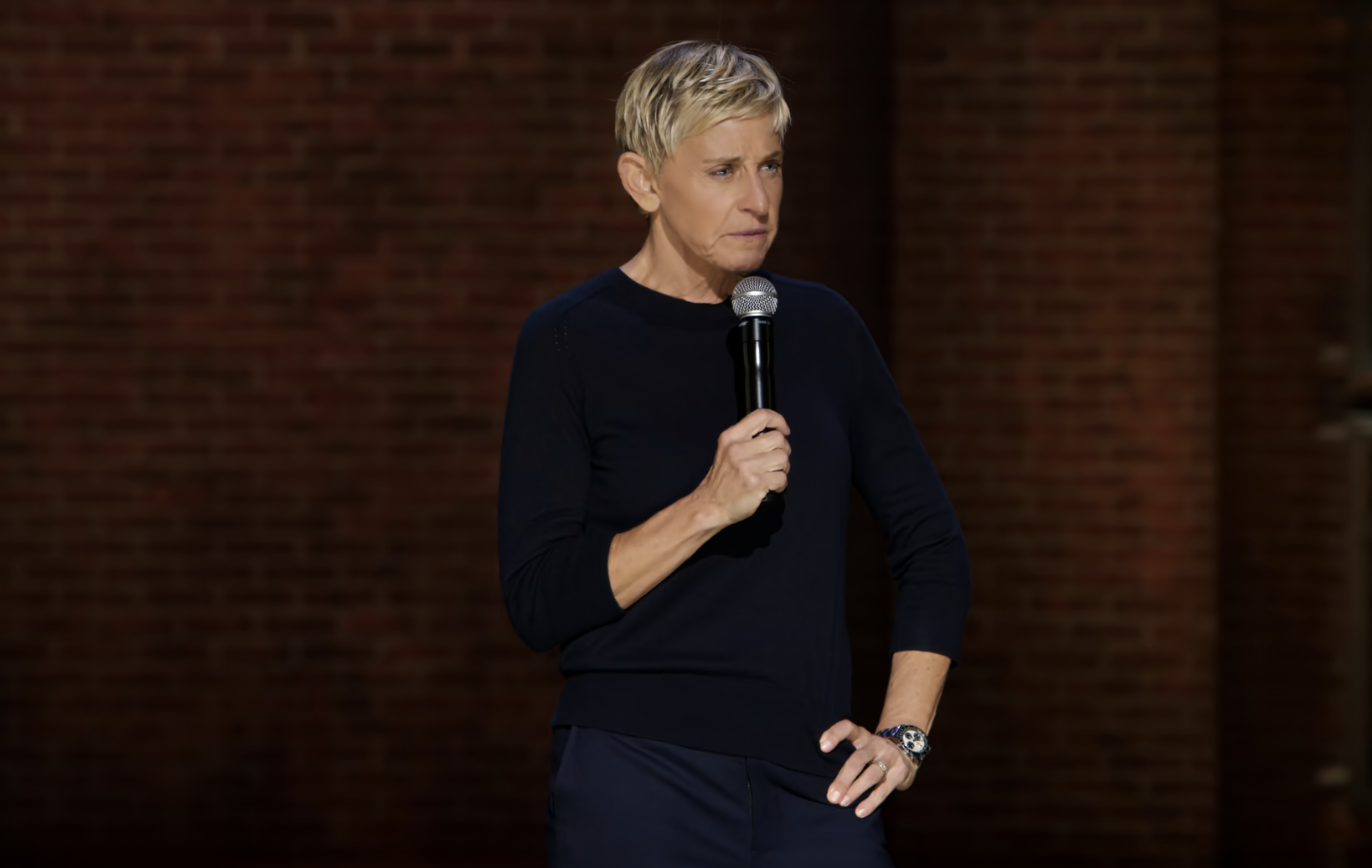 Ellen DeGeneres stands on stage with a microphone in her right hand, wearing a black long-sleeve top and dark pants. She appears to be mid-speech