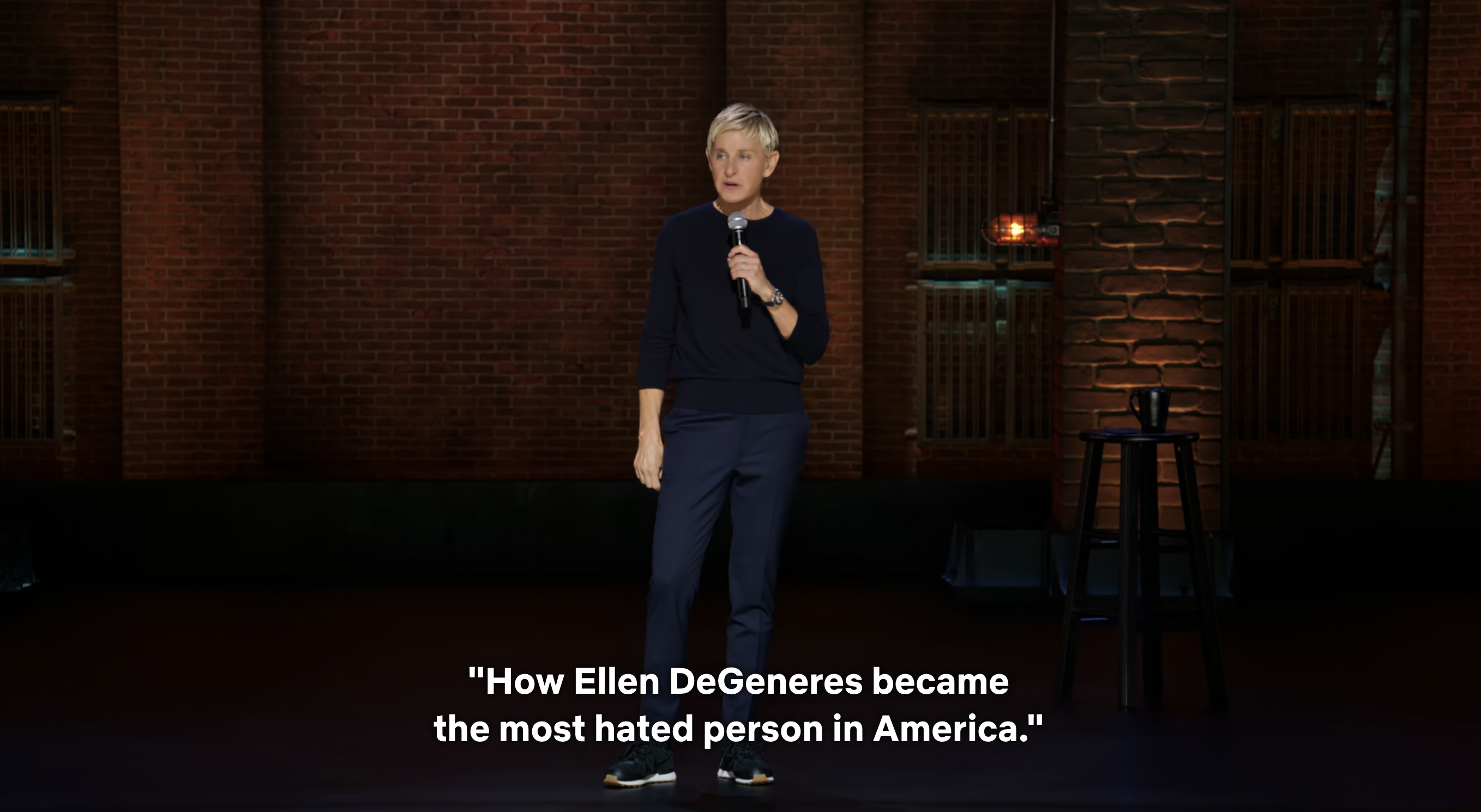 Ellen DeGeneres stands on stage during a stand-up comedy show, holding a microphone. Text below reads, &quot;How Ellen DeGeneres became the most hated person in America&quot;