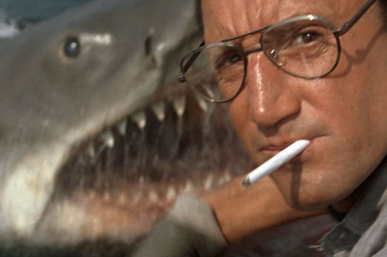 A man with glasses and a cigarette in his mouth looks concerned as a large shark with sharp teeth looms behind him