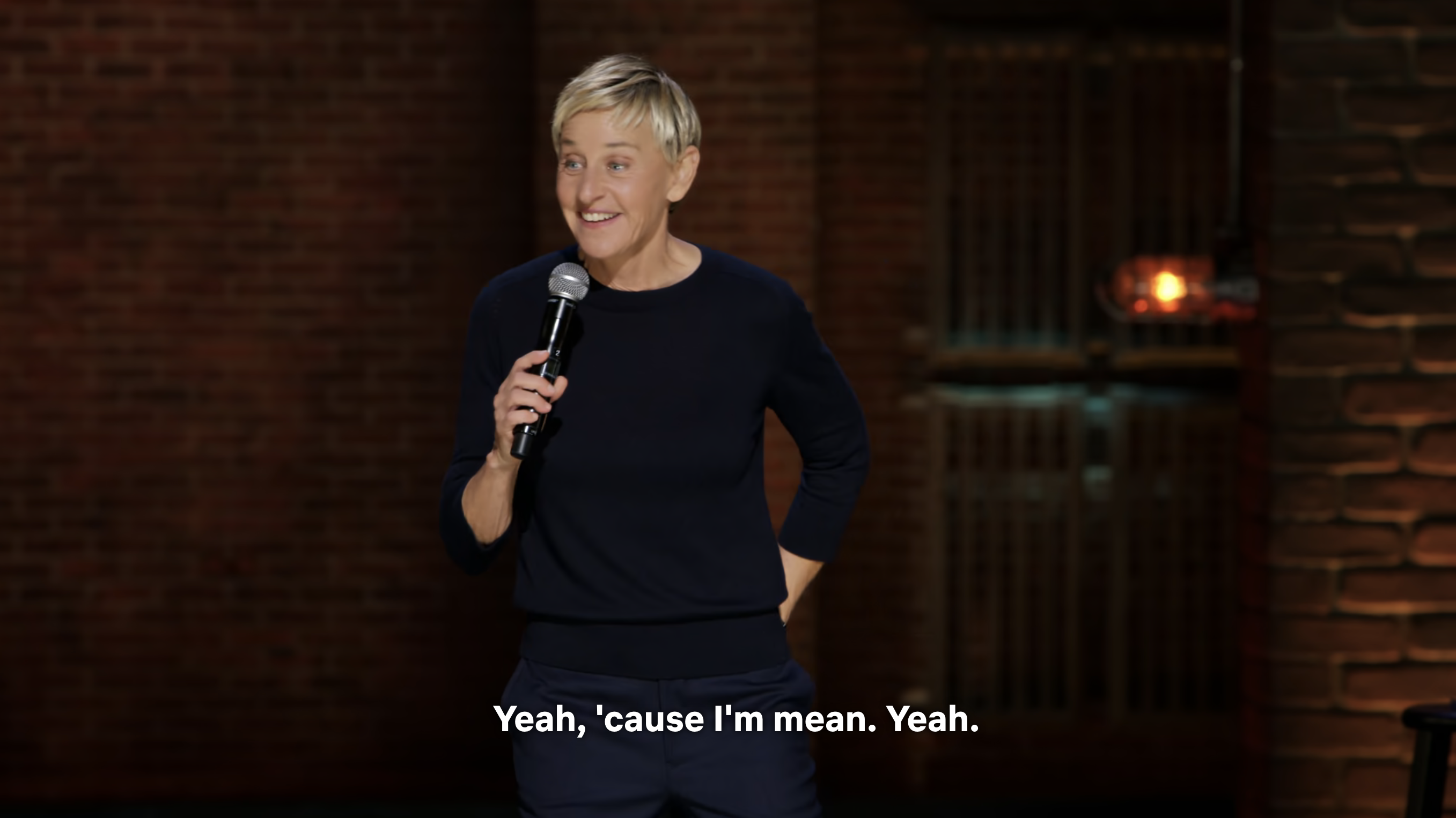 Ellen DeGeneres wearing a simple outfit, performs stand-up comedy on stage, smiling and holding a microphone. Subtitles read: &quot;Yeah, &#x27;cause I&#x27;m mean. Yeah.&quot;