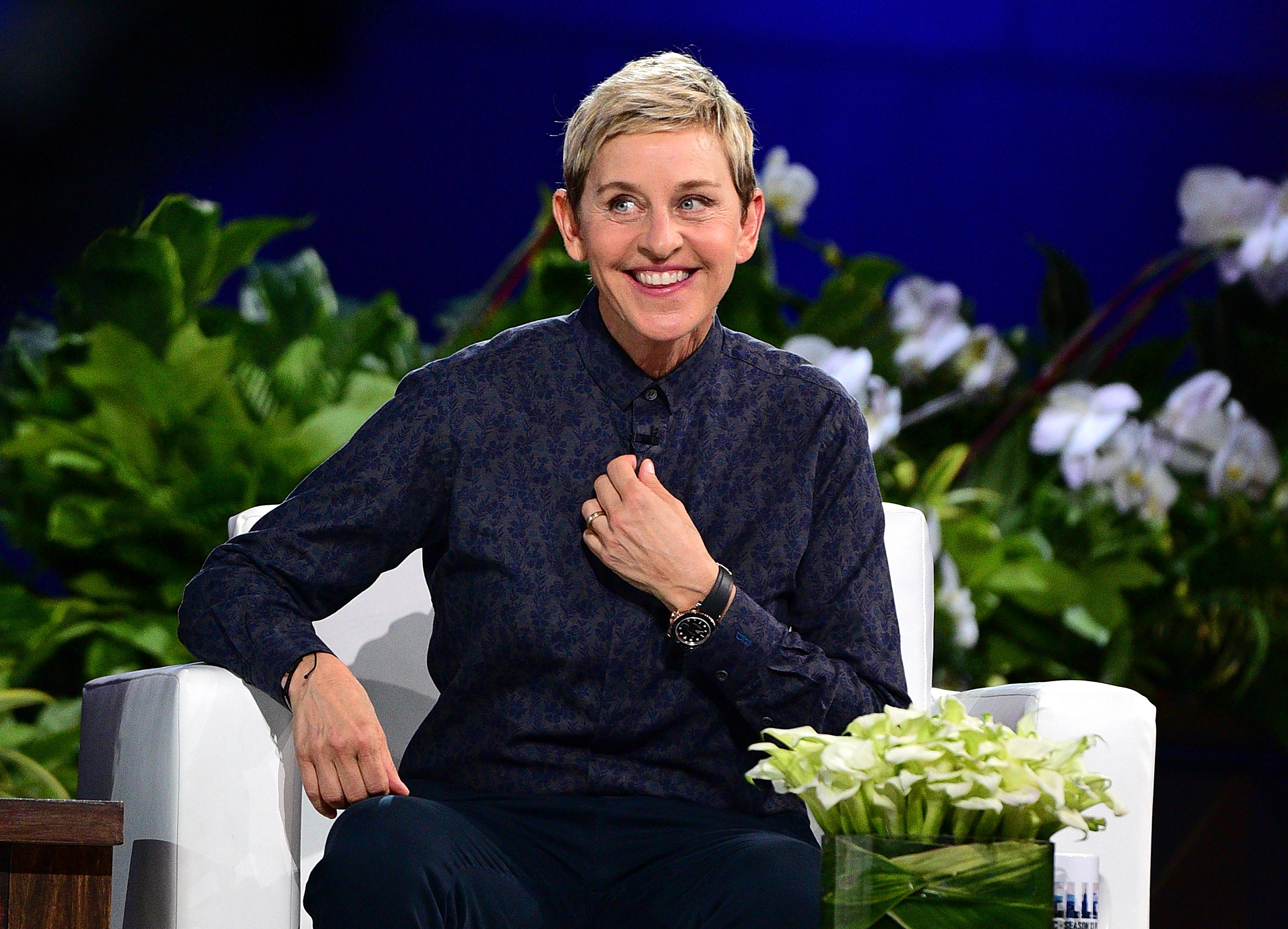 Ellen DeGeneres sits in a white chair on a talk show set, wearing a buttoned-up dark shirt. She smiles and adjusts her collar