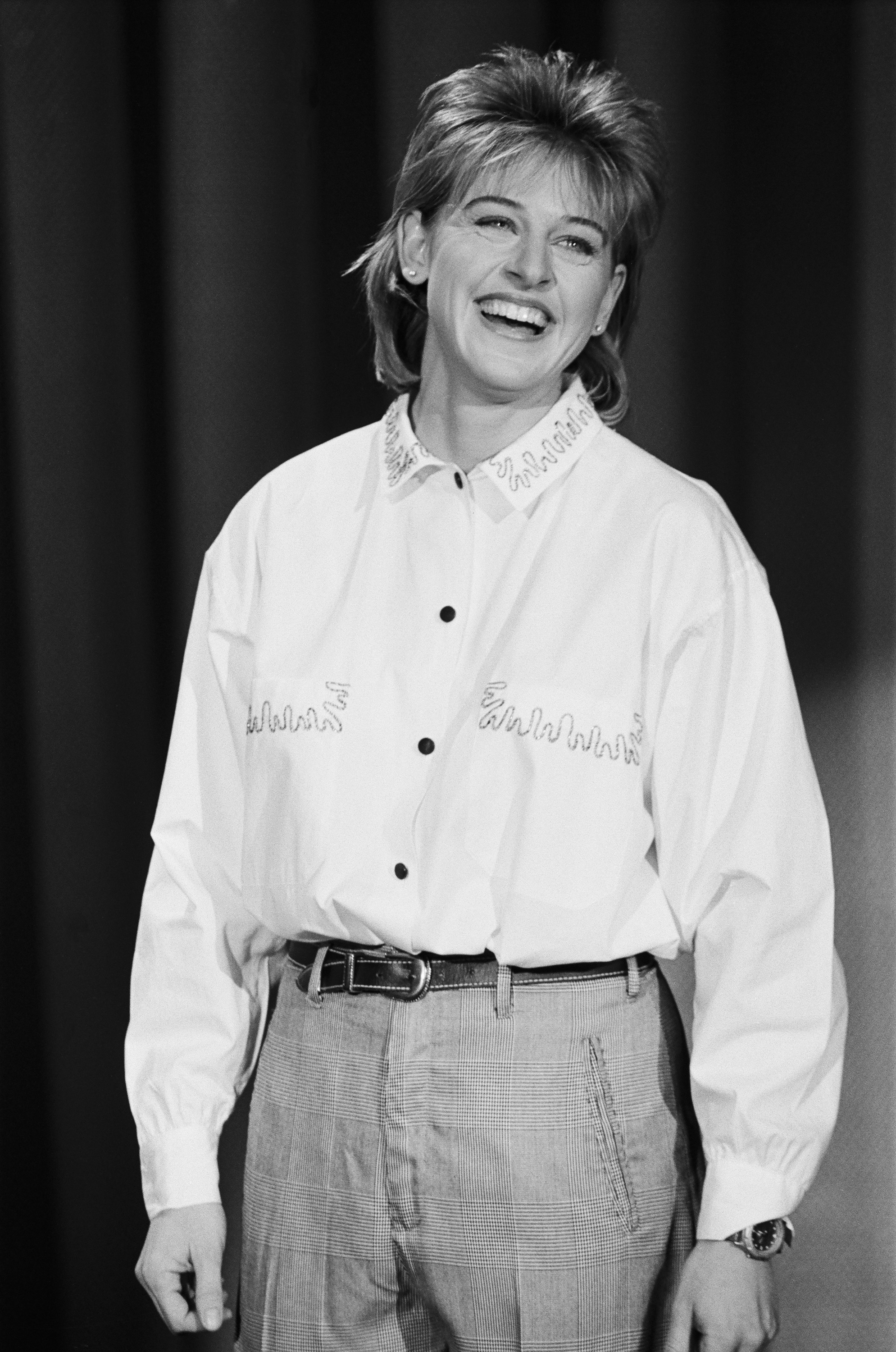 Black and white photo of Ellen DeGeneres smiling and wearing a buttoned-up shirt and high-waisted pants