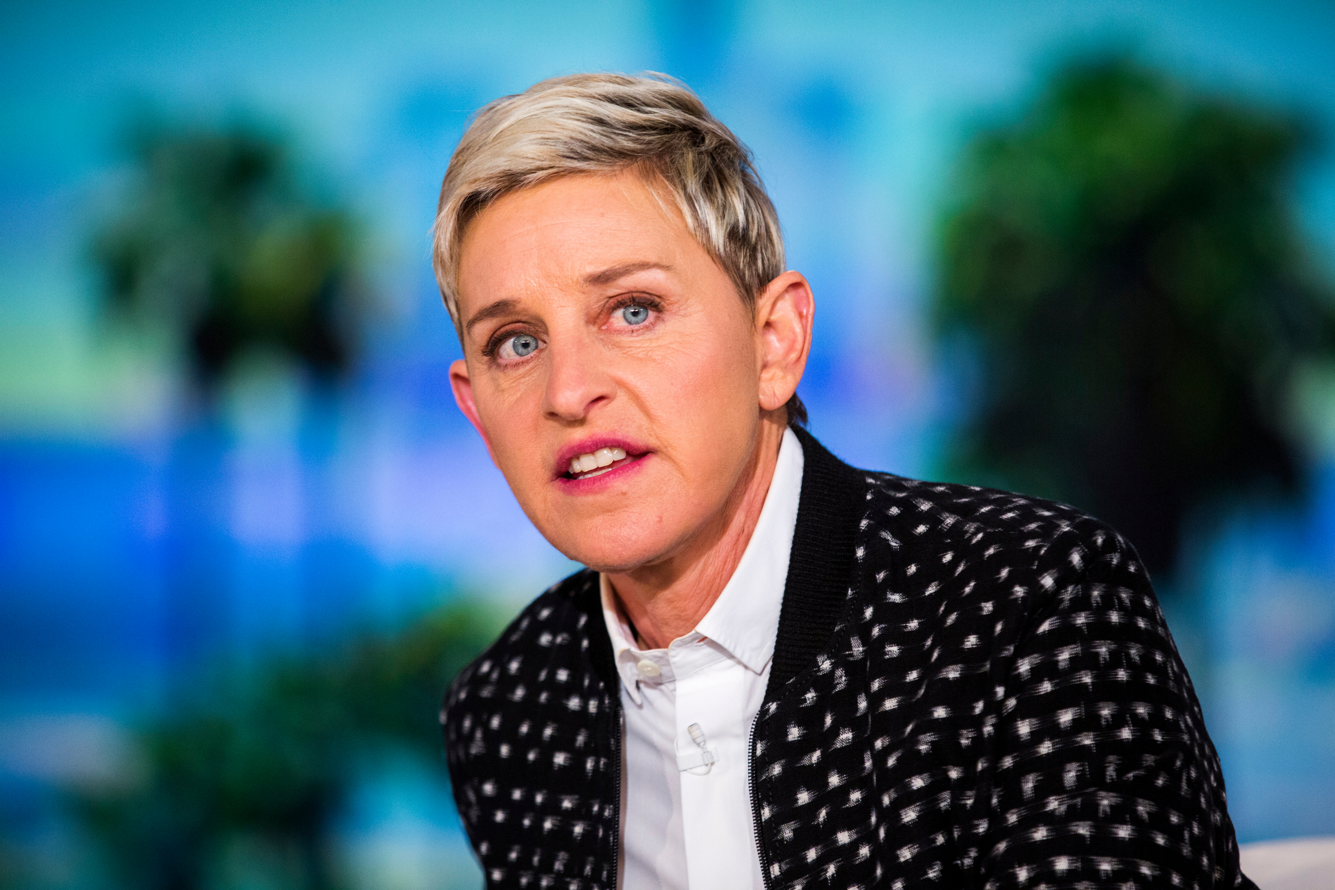 Ellen DeGeneres speaks during an interview. She is wearing a patterned blazer over a shirt. The background is blurred with hints of greenery