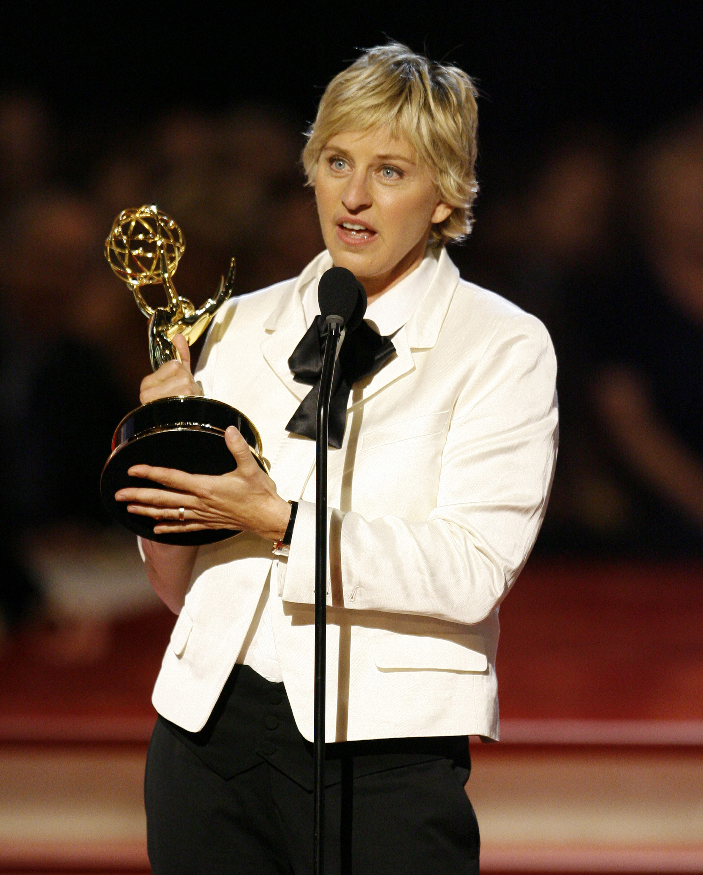 Ellen DeGeneres holds an Emmy Award and speaks into a microphone, wearing a blazer and bow tie