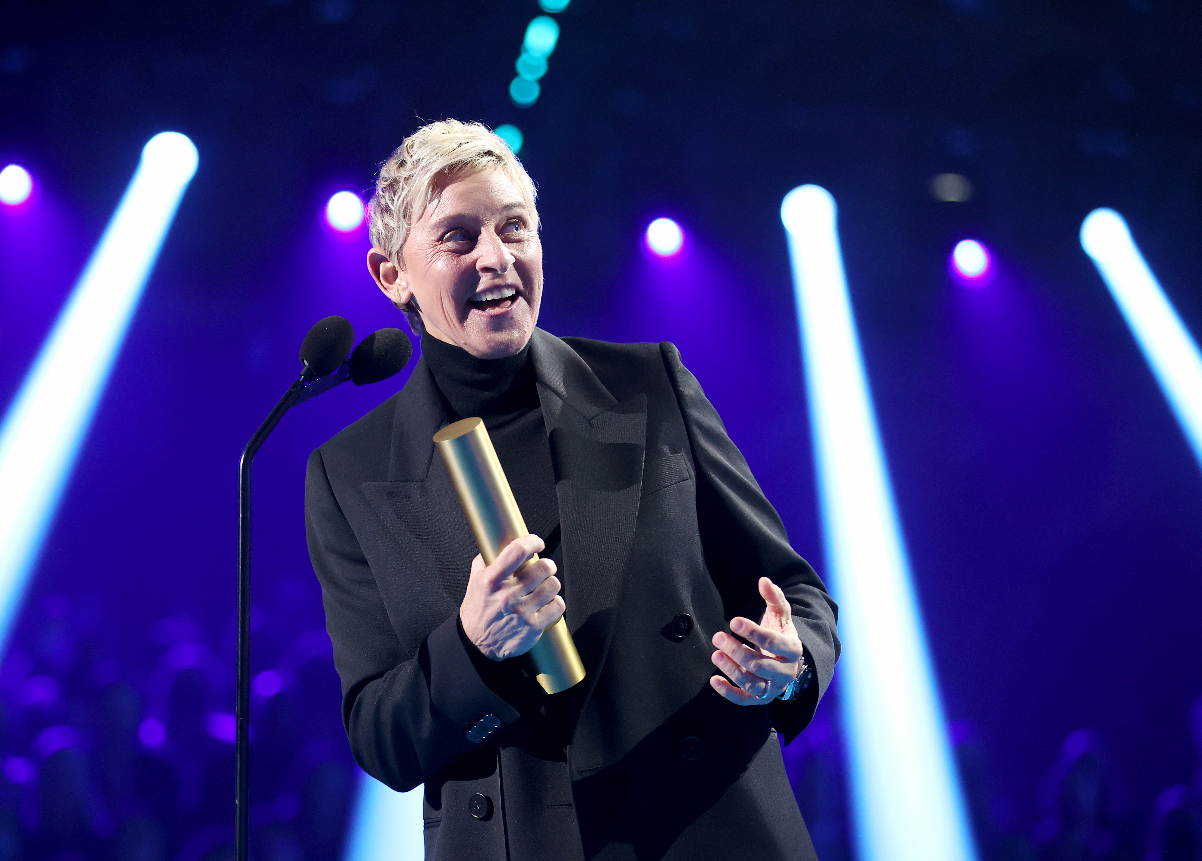 Ellen DeGeneres holding a gold award and speaking on stage at a televised event. She is dressed in a dark suit