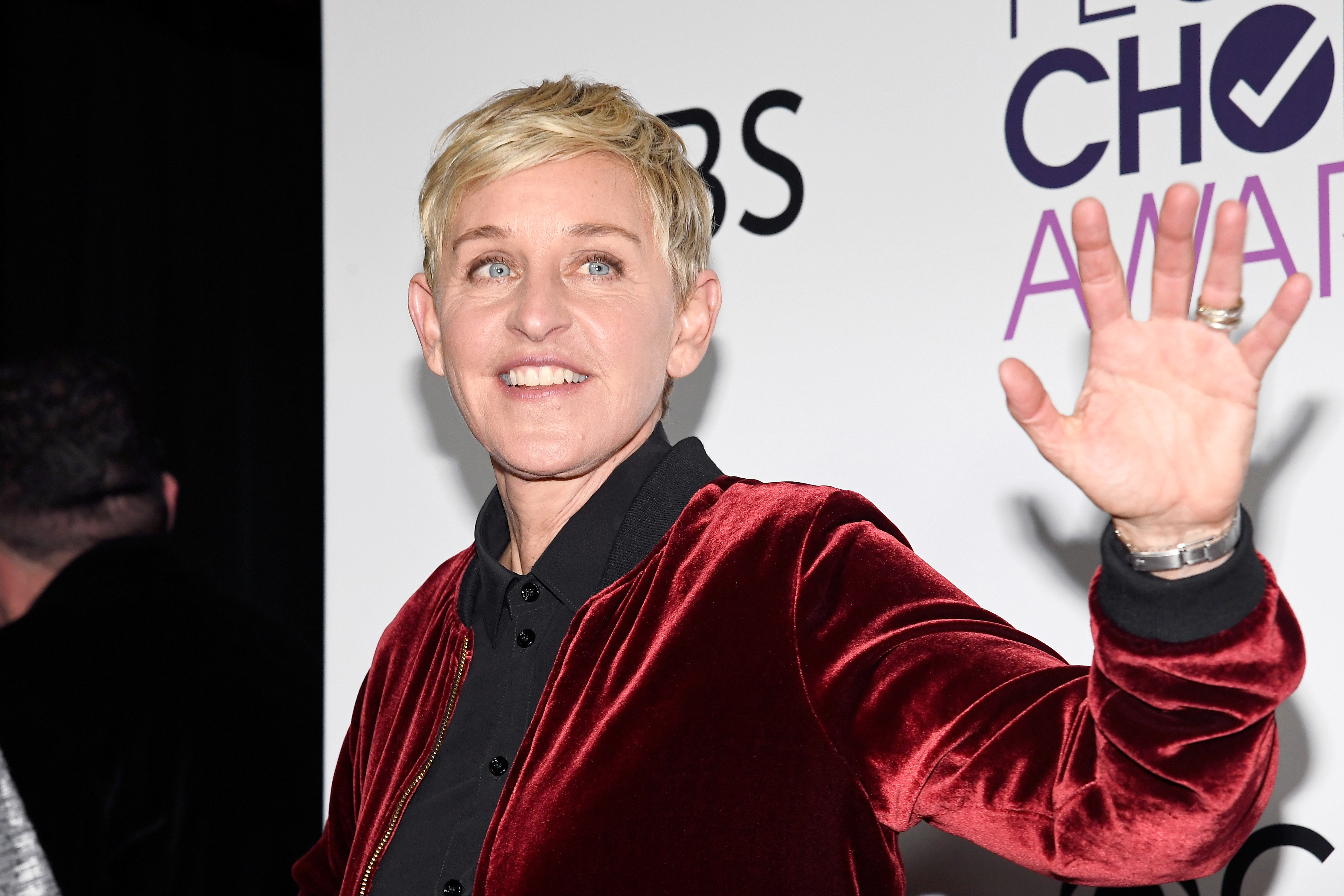 Ellen DeGeneres waves at the People’s Choice Awards, smiling and wearing a velvet jacket over a dark shirt