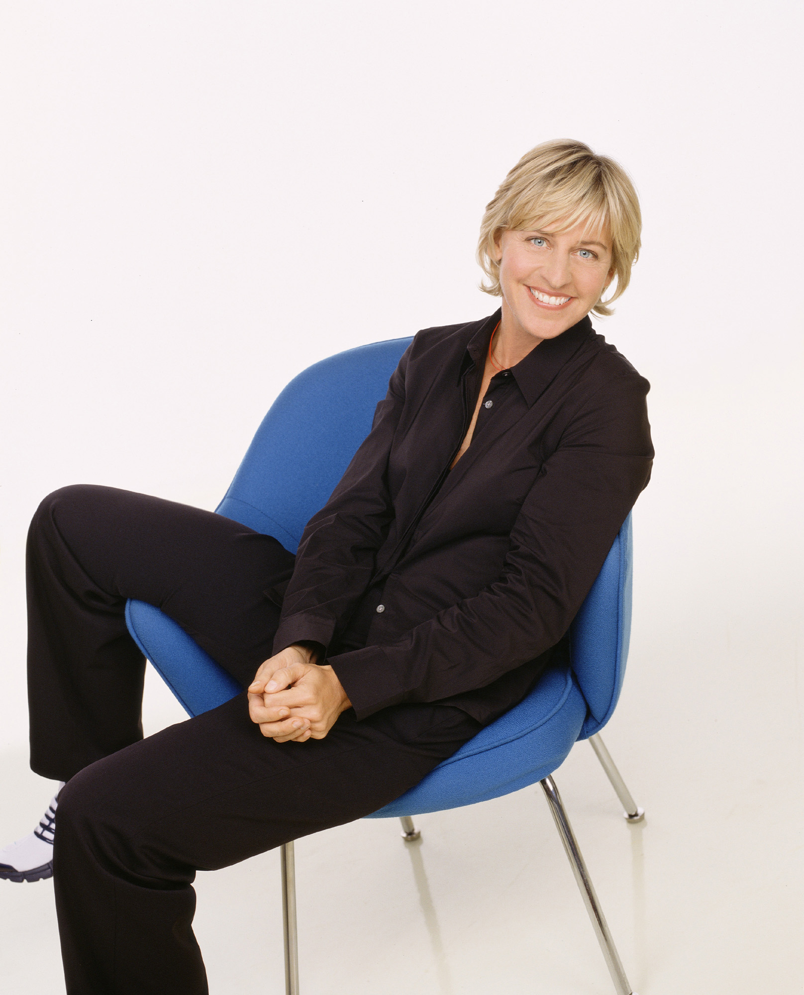 Ellen DeGeneres sits relaxed on a chair, wearing a button-up shirt and pants, smiling at the camera