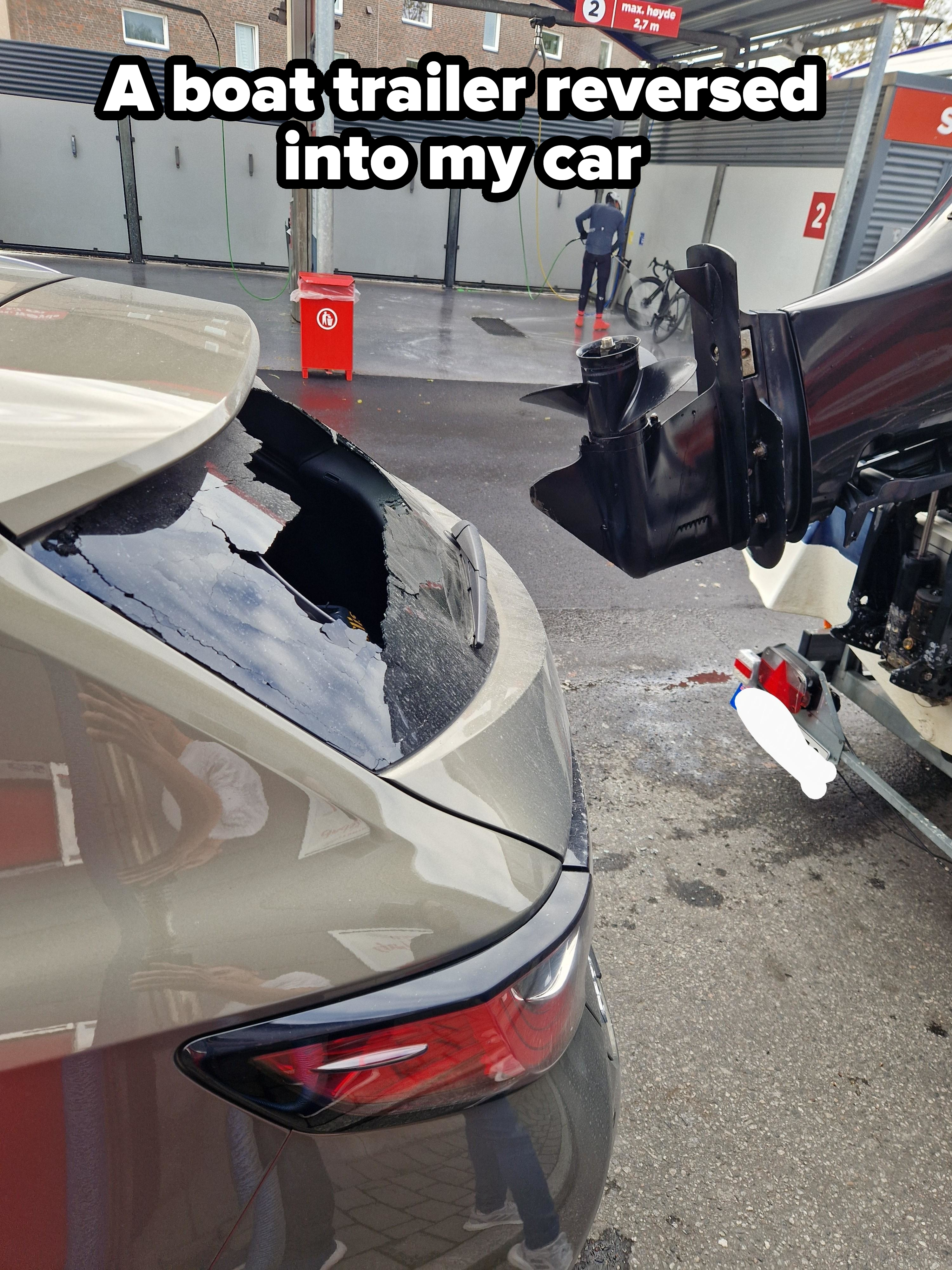 A car with a broken rear windshield is shown at a car wash area, where a boat motor appears to have crashed into it. No people are named in the image