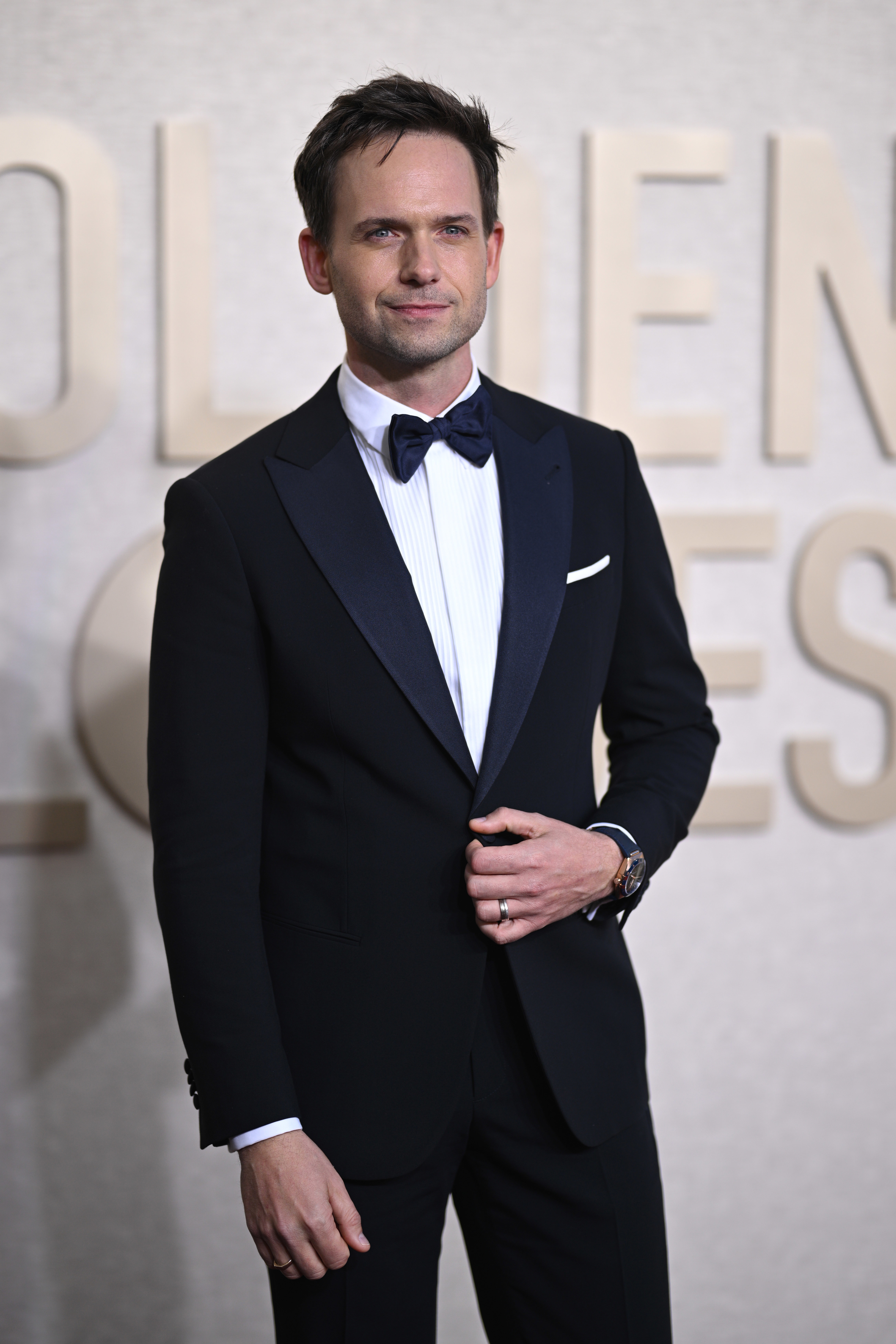 Patrick J. Adams in a classic black tuxedo with a white dress shirt, black bow tie, and wristwatch, posing on the red carpet