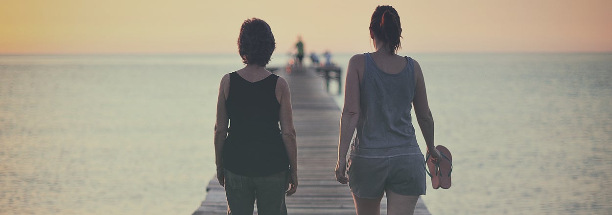 Two people, seen from behind, walking barefoot on a wooden pier towards the horizon. The person on the right holds sandals in their hand