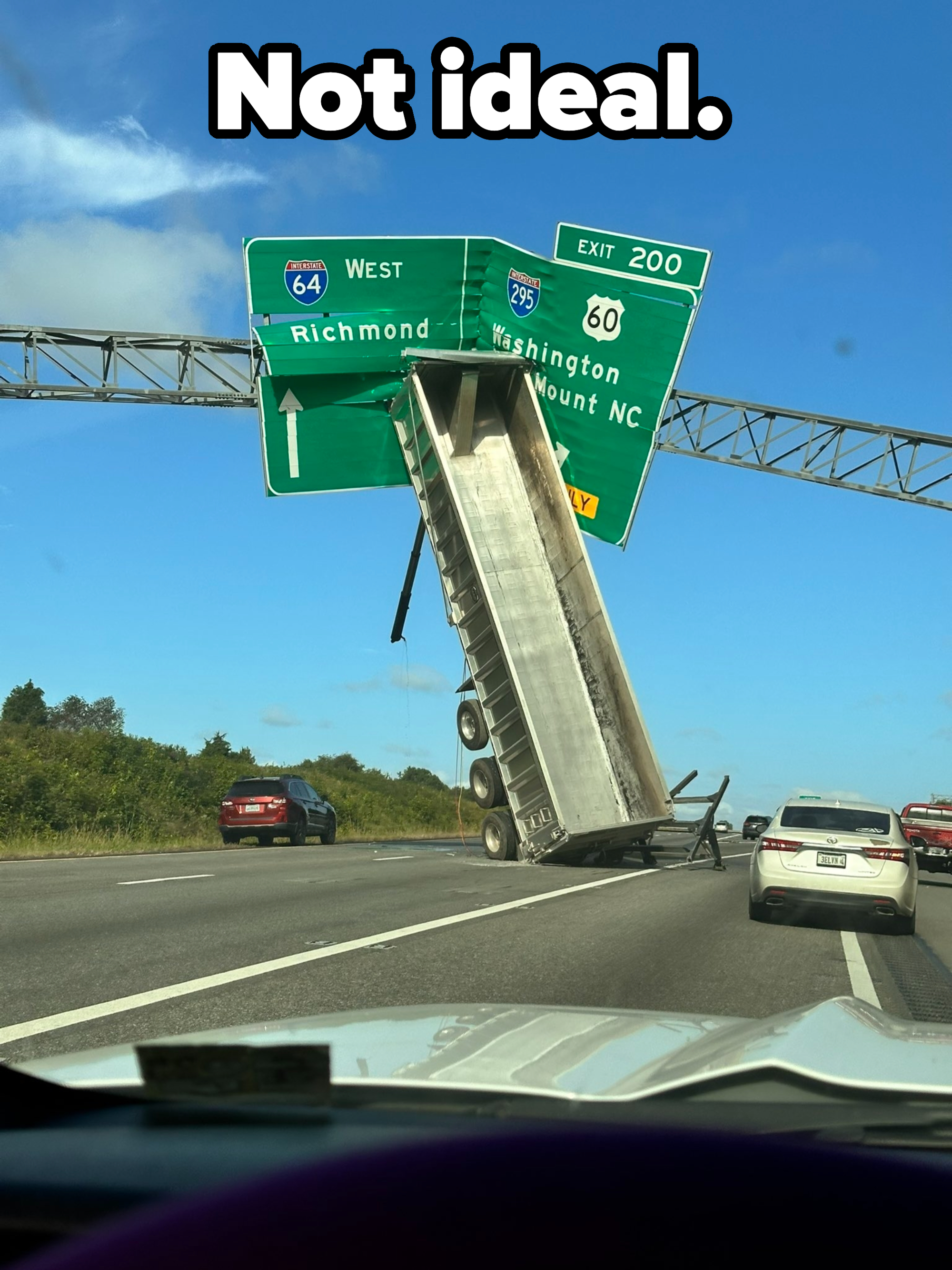 A truck&#x27;s trailer is lifted upright, stuck on a highway exit sign over an interstate, causing traffic to slow down around it