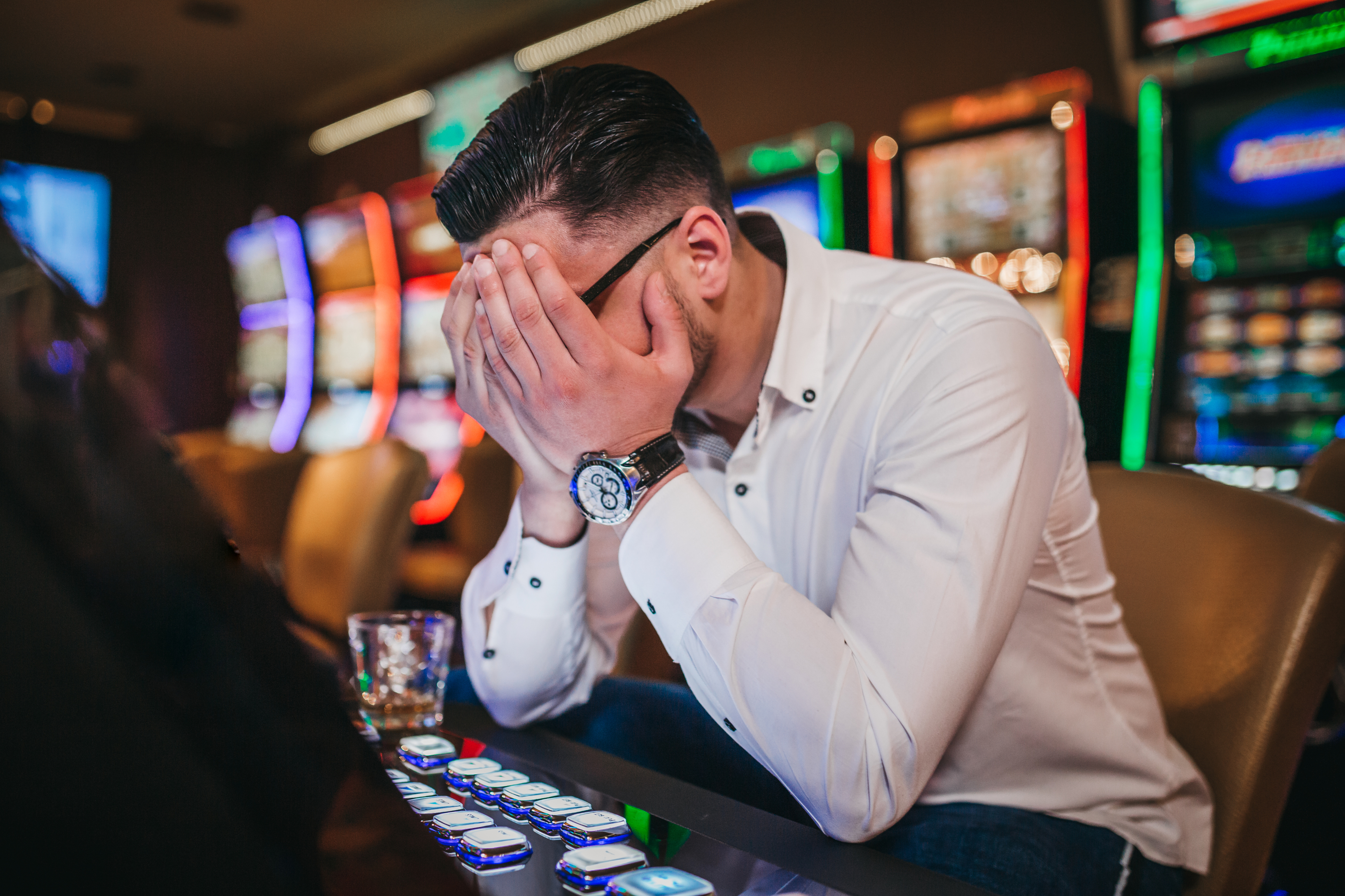 A man sits at a casino slot machine with his face buried in his hands, expressing frustration or distress. A drink is placed on the table beside him