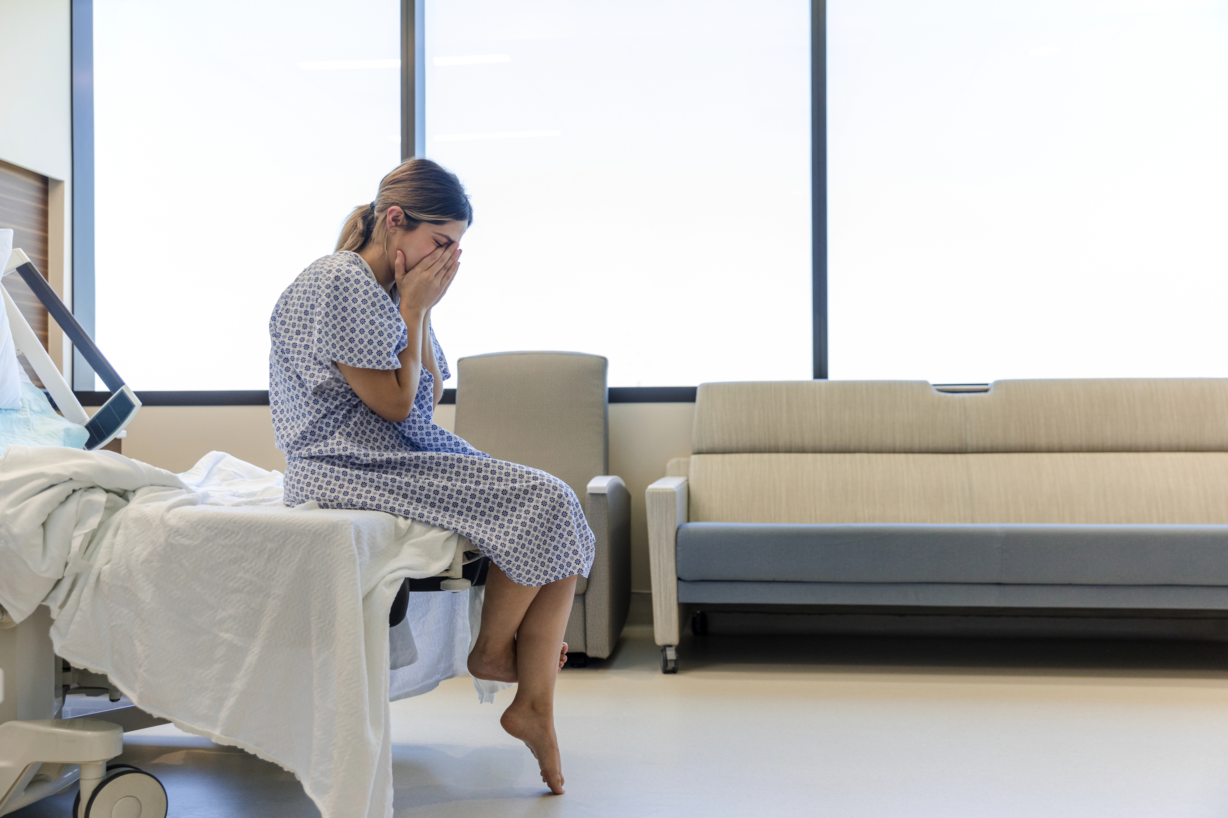 A woman in a hospital gown sits at the edge of a bed with her feet touching the floor, covering her face with her hands in distress