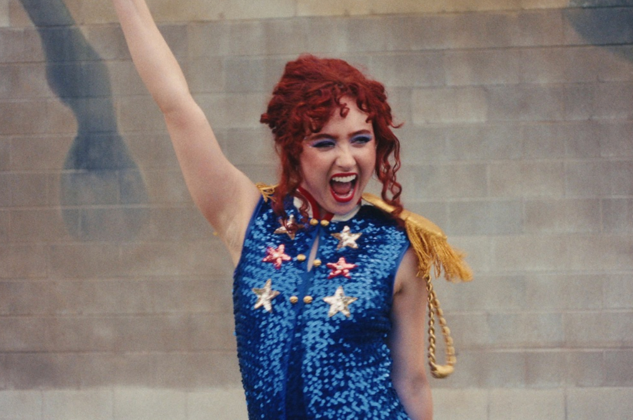 A woman with curly red hair passionately raises one arm while wearing a sequined outfit adorned with star patterns and gold fringe on the shoulders