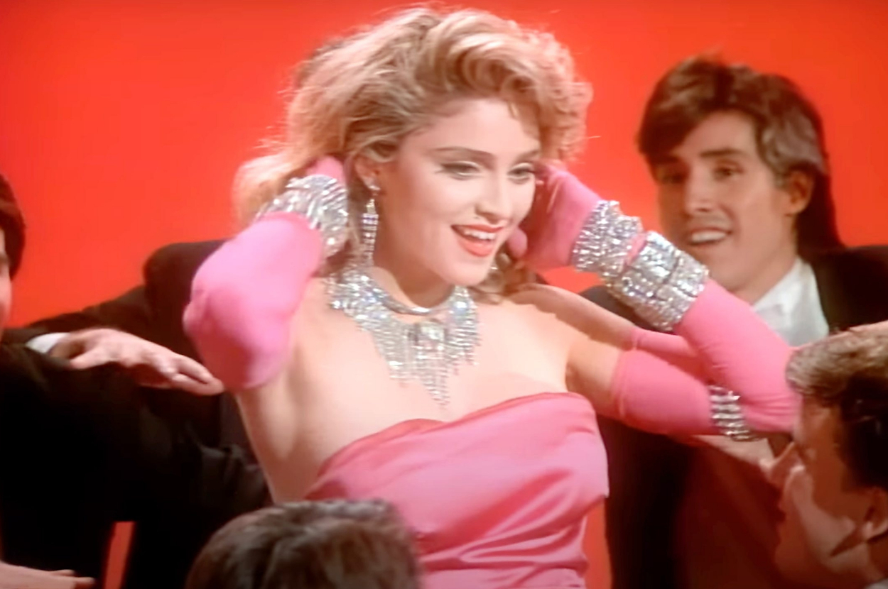 Madonna, in glamorous 1980s attire adorned with sparkling jewelry, is surrounded by people as she puts her hands behind her head against a red backdrop