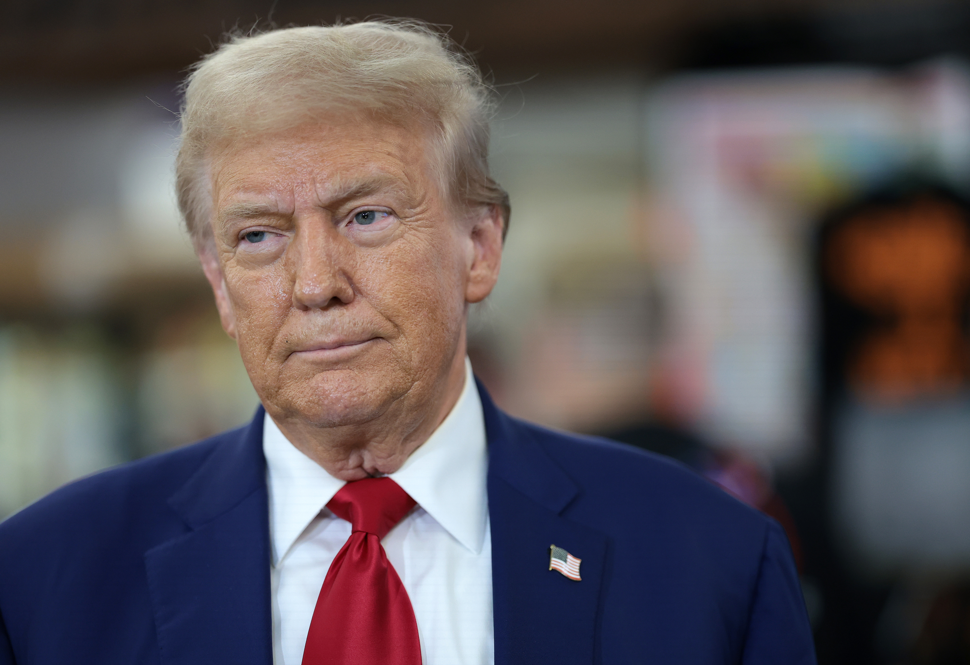 A serious Donald Trump wearing a suit with a red tie and an American flag pin stands indoors