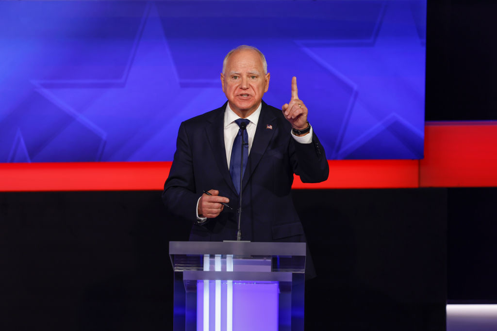 A politician wearing a suit stands at a clear podium, speaking and pointing upward, with a background featuring stars and stripes