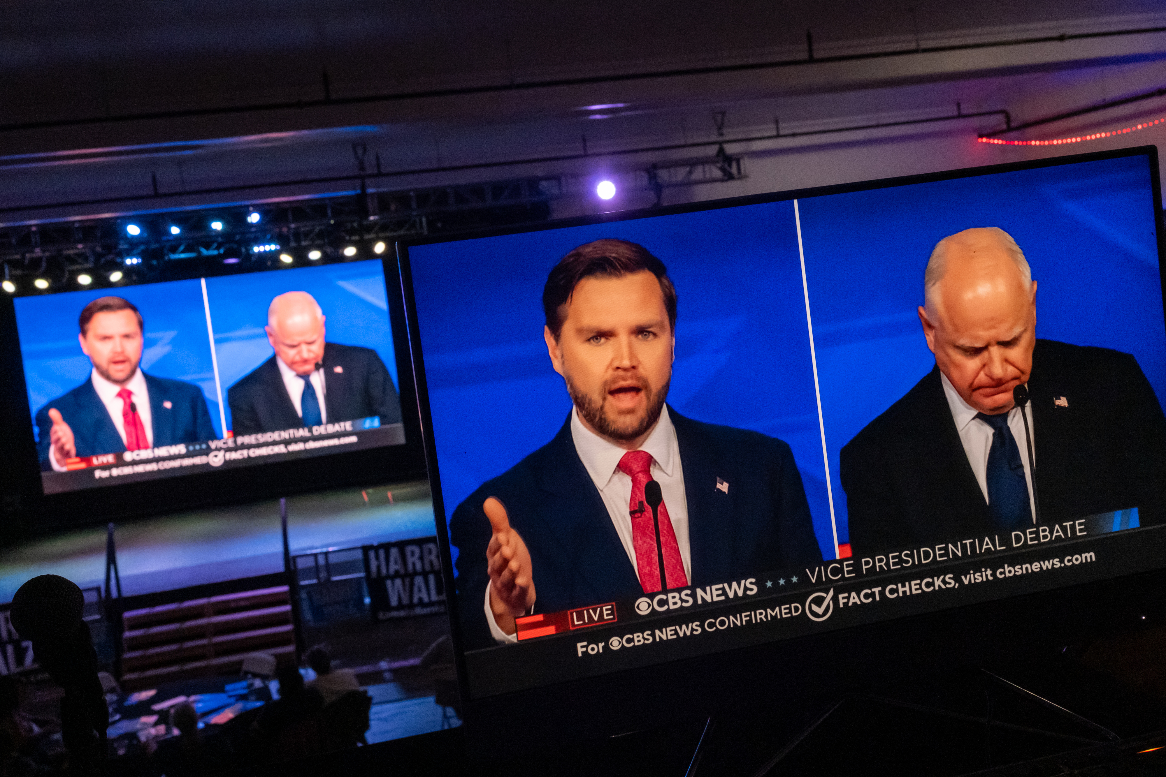 Two men, J.D. Vance and Tim Ryan, are shown on large screens at a vice-presidential debate on CBS News. Ryan is making a speech while Vance looks down