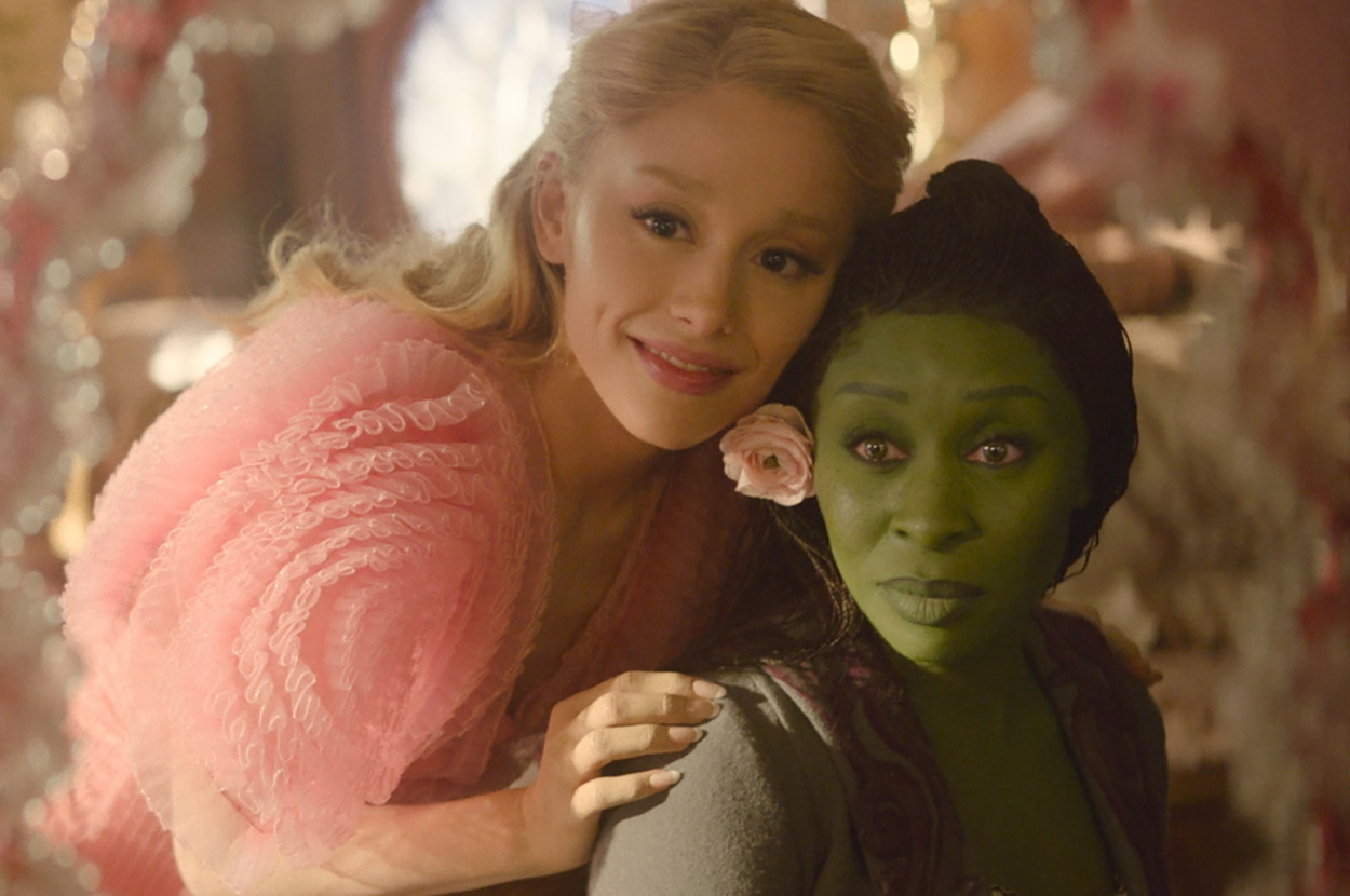 Ariana Grande, dressed in a pink, frilly outfit, is smiling next to Cynthia Erivo, who has green skin and a flower in her hair