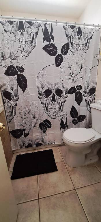 A bathroom with a shower curtain featuring a black and white skull and floral design. There is a white toilet and a black rug on the floor