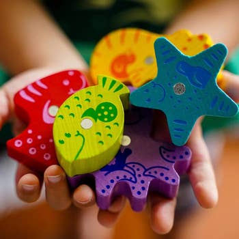 Hands holding colorful wooden toy sea creatures: a starfish, fish, and squid. Image promotes children's educational toys for sale