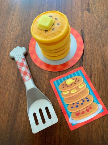 Plastic toy pancakes with a spatula on a table. A card showing three pancakes with different toppings is next to the toy