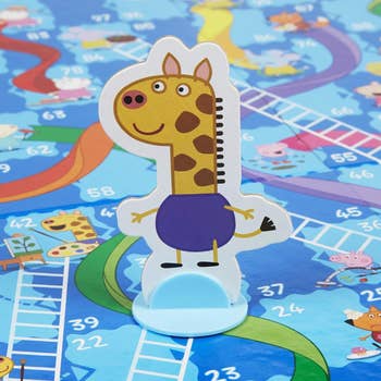 A toy figure of a smiling giraffe in purple shorts is placed on a colorful board game featuring ladders and slides, numbers, and cartoon characters