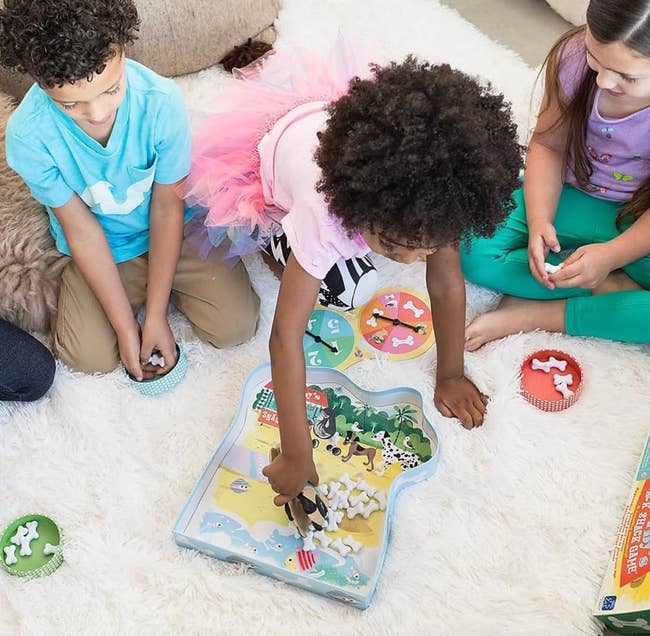 Three children play with a dinosaur-themed board game on a fluffy white rug