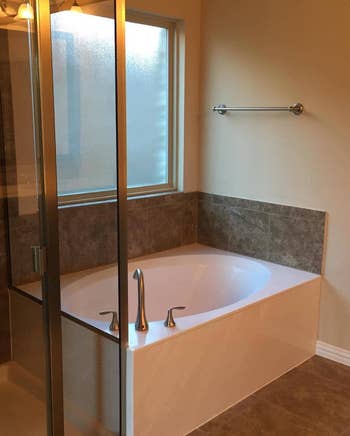 A corner bathtub with a glass shower partition and a frosted window above in a tiled bathroom. No persons are depicted