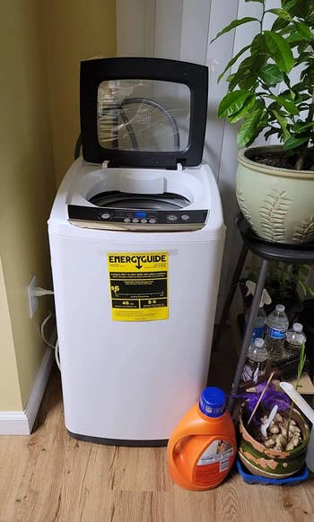 A top-loading washing machine with an open lid, placed next to a plant and a shelf with supplies. An energy guide label is visible on the front