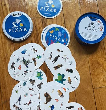 Spot It! Pixar game cards scattered on a wooden floor with game rule booklets and a tin container